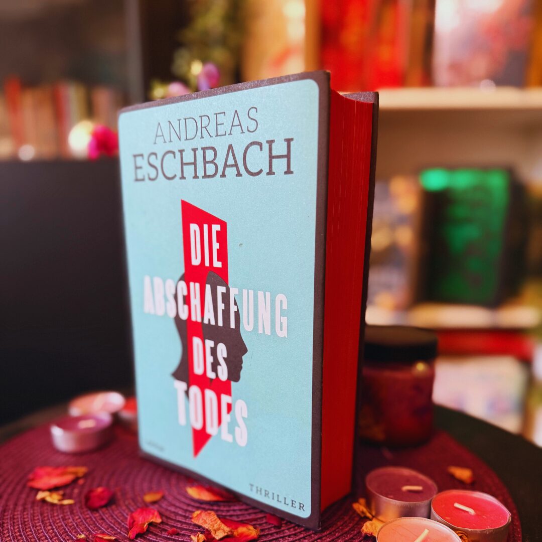 Das Bild zeigt ein Buch mit dem Titel „Die Abschaffung des Todes“ von Andreas Eschbach. Der Buchumschlag ist hellblau mit einem roten Längsstreifen in der Mitte, der die Silhouette des Profils einer Person darstellt. Der Titel steht in fetter weißer Schrift über dem roten Streifen. Der Name des Autors, Andreas Eschbach, steht oben in schwarzen Buchstaben. Das Wort „Thriller“ ist in der unteren rechten Ecke des Covers aufgedruckt. Das Buch steht aufrecht auf einer mit einer runden violetten Matte geschmückten Fläche, umgeben von kleinen Kerzen und verstreuten getrockneten Blütenblättern. Im Hintergrund sind verschwommene Bücherregale mit verschiedenen Büchern zu sehen.