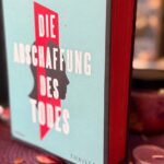 Das Bild zeigt ein Buch mit dem Titel „Die Abschaffung des Todes“ von Andreas Eschbach. Der Buchumschlag ist hellblau mit einem roten Längsstreifen in der Mitte, der die Silhouette des Profils einer Person darstellt. Der Titel steht in fetter weißer Schrift über dem roten Streifen. Der Name des Autors, Andreas Eschbach, steht oben in schwarzen Buchstaben. Das Wort „Thriller“ ist in der unteren rechten Ecke des Covers aufgedruckt. Das Buch steht aufrecht auf einer mit einer runden violetten Matte geschmückten Fläche, umgeben von kleinen Kerzen und verstreuten getrockneten Blütenblättern. Im Hintergrund sind verschwommene Bücherregale mit verschiedenen Büchern zu sehen.