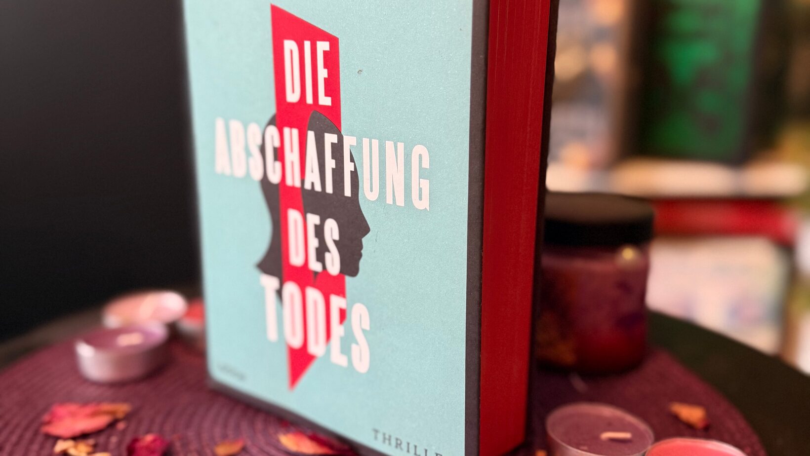 Das Bild zeigt ein Buch mit dem Titel „Die Abschaffung des Todes“ von Andreas Eschbach. Der Buchumschlag ist hellblau mit einem roten Längsstreifen in der Mitte, der die Silhouette des Profils einer Person darstellt. Der Titel steht in fetter weißer Schrift über dem roten Streifen. Der Name des Autors, Andreas Eschbach, steht oben in schwarzen Buchstaben. Das Wort „Thriller“ ist in der unteren rechten Ecke des Covers aufgedruckt. Das Buch steht aufrecht auf einer mit einer runden violetten Matte geschmückten Fläche, umgeben von kleinen Kerzen und verstreuten getrockneten Blütenblättern. Im Hintergrund sind verschwommene Bücherregale mit verschiedenen Büchern zu sehen.