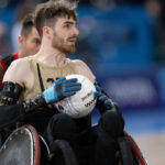 Das Bild zeigt einen männlichen Athleten, der an einem Rollstuhl-Rugbyspiel teilnimmt. Er trägt ein ärmelloses goldenes Trikot mit der Nummer 23 darauf. Der Sportler hat kurze braune Haare und einen Bart. Mit beiden Händen hält er einen weißen Rugbyball, auf dem Ball sind die Worte „molten“ und „WWR“ aufgedruckt. Der Athlet trägt schwarze Handschuhe und hat Schutzausrüstung an den Armen. Hinter ihm ist teilweise ein weiterer Athlet zu sehen, der ein schwarz-rotes Trikot trägt. Die Kulisse scheint eine Indoor-Sportarena mit unscharfem Hintergrund zu sein.