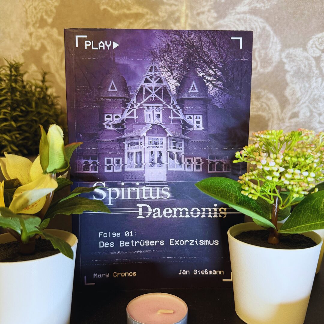 Das Bild zeigt ein Buchcover mit dem Titel "Spiritus Daemonis". Im Hintergrund ist ein großes, altes Haus abgebildet, das in einem düsteren, violetten Farbton gehalten ist. Oben links steht "PLAY" in weißen Buchstaben. Unter dem Titel steht "Folge 01: Des Betrügers Exorzismus". Die Autoren des Buches sind Mary Cronos und Jan Gießmann, deren Namen unten auf dem Cover stehen. Vor dem Buch stehen zwei kleine Pflanzen in weißen Töpfen und eine kleine, runde Kerze. Der Hintergrund des Bildes ist eine gemusterte Tapete.