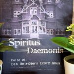 Das Bild zeigt ein Buchcover mit dem Titel "Spiritus Daemonis". Im Hintergrund ist ein großes, altes Haus abgebildet, das in einem düsteren, violetten Farbton gehalten ist. Oben links steht "PLAY" in weißen Buchstaben. Unter dem Titel steht "Folge 01: Des Betrügers Exorzismus". Die Autoren des Buches sind Mary Cronos und Jan Gießmann, deren Namen unten auf dem Cover stehen. Vor dem Buch stehen zwei kleine Pflanzen in weißen Töpfen und eine kleine, runde Kerze. Der Hintergrund des Bildes ist eine gemusterte Tapete.