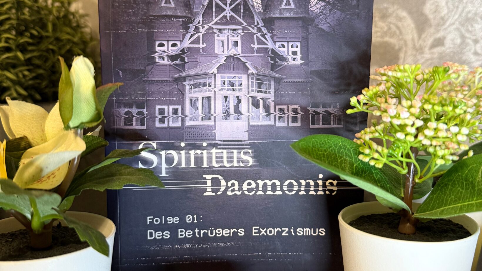 Das Bild zeigt ein Buchcover mit dem Titel "Spiritus Daemonis". Im Hintergrund ist ein großes, altes Haus abgebildet, das in einem düsteren, violetten Farbton gehalten ist. Oben links steht "PLAY" in weißen Buchstaben. Unter dem Titel steht "Folge 01: Des Betrügers Exorzismus". Die Autoren des Buches sind Mary Cronos und Jan Gießmann, deren Namen unten auf dem Cover stehen. Vor dem Buch stehen zwei kleine Pflanzen in weißen Töpfen und eine kleine, runde Kerze. Der Hintergrund des Bildes ist eine gemusterte Tapete.