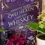 Das Bild zeigt das Cover eines Buches mit dem Titel "Zwei Hexen und ein Whiskey" von Annette Marie. Es ist der dritte Band der Reihe "Guild Codex: Spellbound". Das Cover ist in einem kräftigen Lila gehalten und mit goldenen Verzierungen geschmückt. In der Mitte des Covers ist ein stilisiertes Bild eines Eulenmotivs zu sehen. Um das Buch herum stehen mehrere kleine Pflanzen in weißen Töpfen, darunter eine rote Blume im Vordergrund. Der Hintergrund ist eine beige Tapete mit einem dezenten Muster.