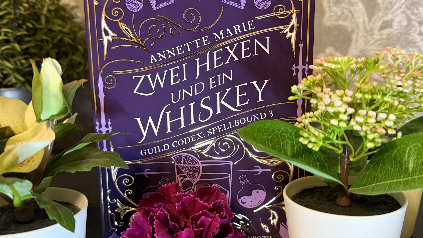 Das Bild zeigt das Cover eines Buches mit dem Titel "Zwei Hexen und ein Whiskey" von Annette Marie. Es ist der dritte Band der Reihe "Guild Codex: Spellbound". Das Cover ist in einem kräftigen Lila gehalten und mit goldenen Verzierungen geschmückt. In der Mitte des Covers ist ein stilisiertes Bild eines Eulenmotivs zu sehen. Um das Buch herum stehen mehrere kleine Pflanzen in weißen Töpfen, darunter eine rote Blume im Vordergrund. Der Hintergrund ist eine beige Tapete mit einem dezenten Muster.