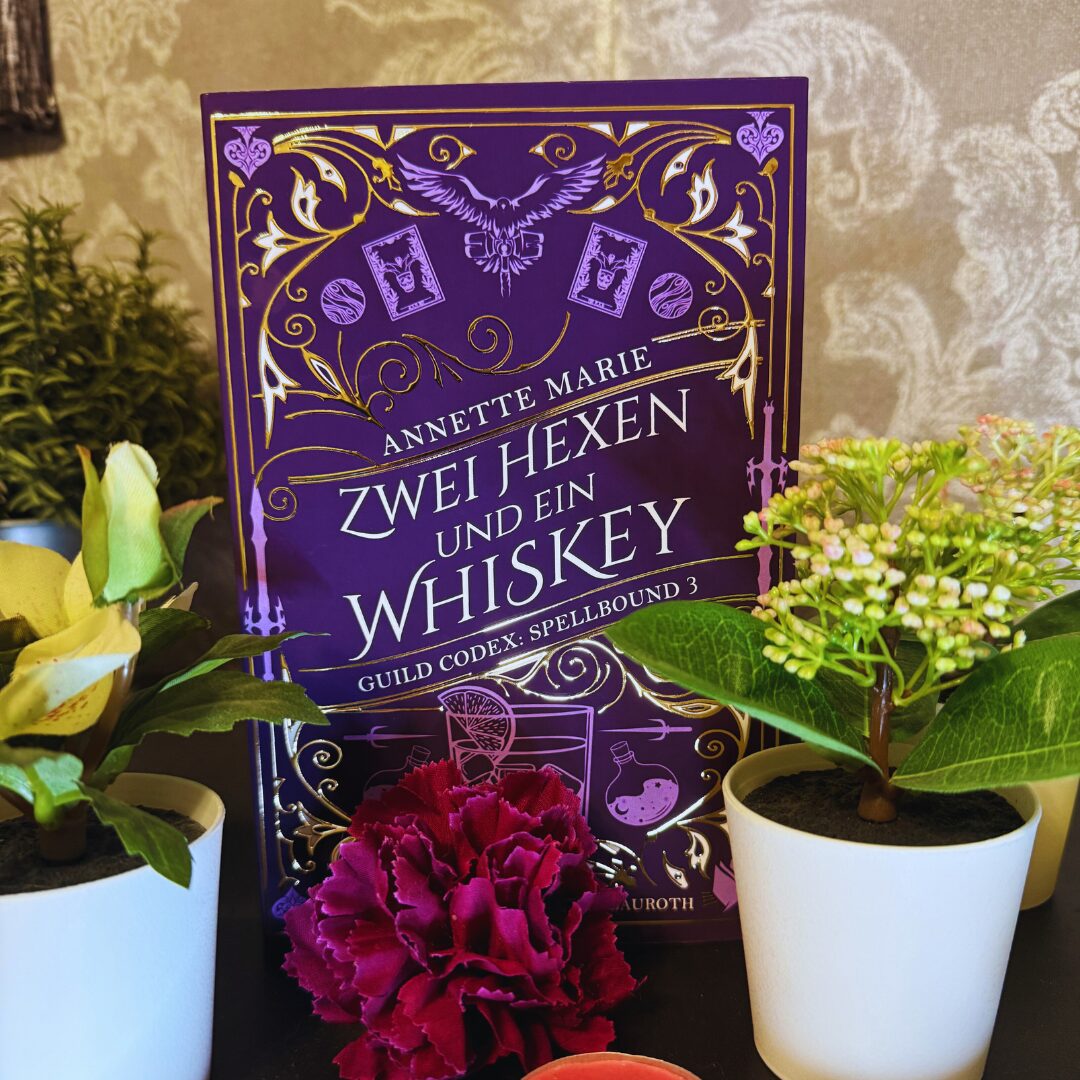 Das Bild zeigt das Cover eines Buches mit dem Titel "Zwei Hexen und ein Whiskey" von Annette Marie. Es ist der dritte Band der Reihe "Guild Codex: Spellbound". Das Cover ist in einem kräftigen Lila gehalten und mit goldenen Verzierungen geschmückt. In der Mitte des Covers ist ein stilisiertes Bild eines Eulenmotivs zu sehen. Um das Buch herum stehen mehrere kleine Pflanzen in weißen Töpfen, darunter eine rote Blume im Vordergrund. Der Hintergrund ist eine beige Tapete mit einem dezenten Muster.