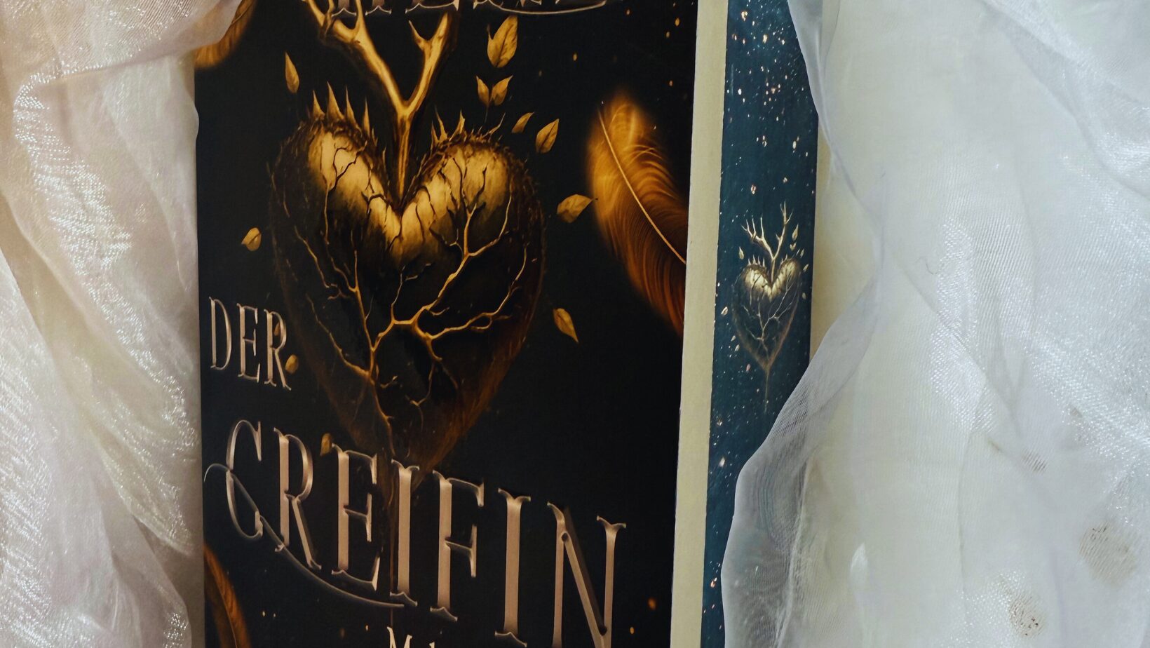 ein Buch mit dem Titel "Das Herz der Greifin: Malinas Rache" von Isabell Bayer. Auf dem Buchcover ist ein goldenes Herz mit Ästen und Blättern abgebildet, umgeben von goldenen Federn. Der Hintergrund ist dunkel gehalten, was das Herz und die Federn hervorhebt. Unten auf dem Cover steht "Lycrow Verlag". Das Buch ist in ein weißes, leicht transparentes Tuch eingewickelt.