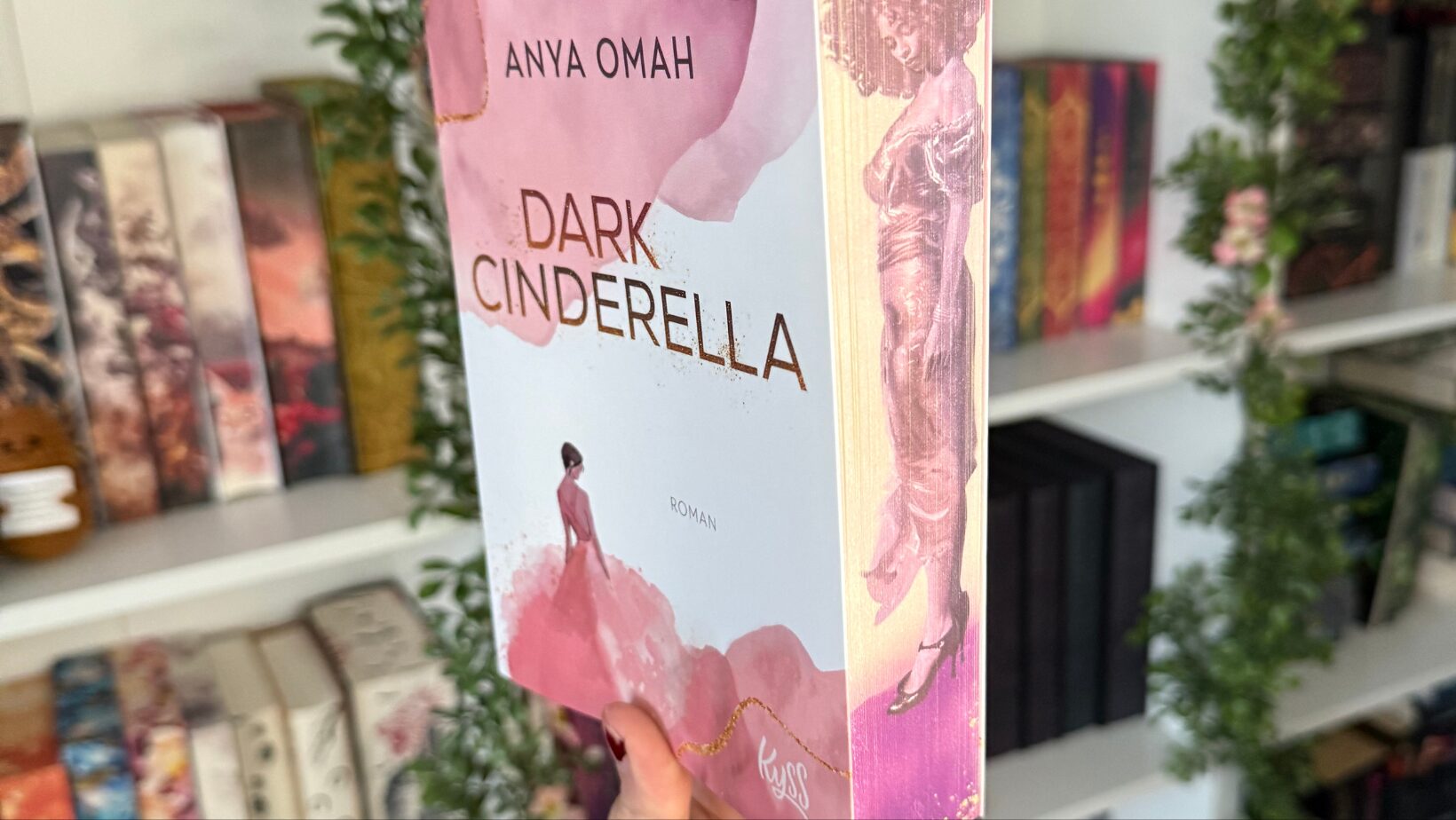 ein Buch, das in einer Hand gehalten wird. Der Titel des Buches ist "Dark Cinderella" und die Autorin ist Anya Omah. Auf dem Buchcover ist eine stilisierte Illustration einer Frau in einem rosa Kleid zu sehen. Oben auf dem Cover befindet sich ein runder Aufkleber mit der Aufschrift "#BookTok Bestseller Autorin". Im Hintergrund sind Bücherregale mit verschiedenen Büchern und grünen Pflanzen zu sehen. Das Buch hat einen rosa-weißen Farbverlauf und der Buchrücken zeigt eine weitere Illustration einer Frau in einem langen Kleid.