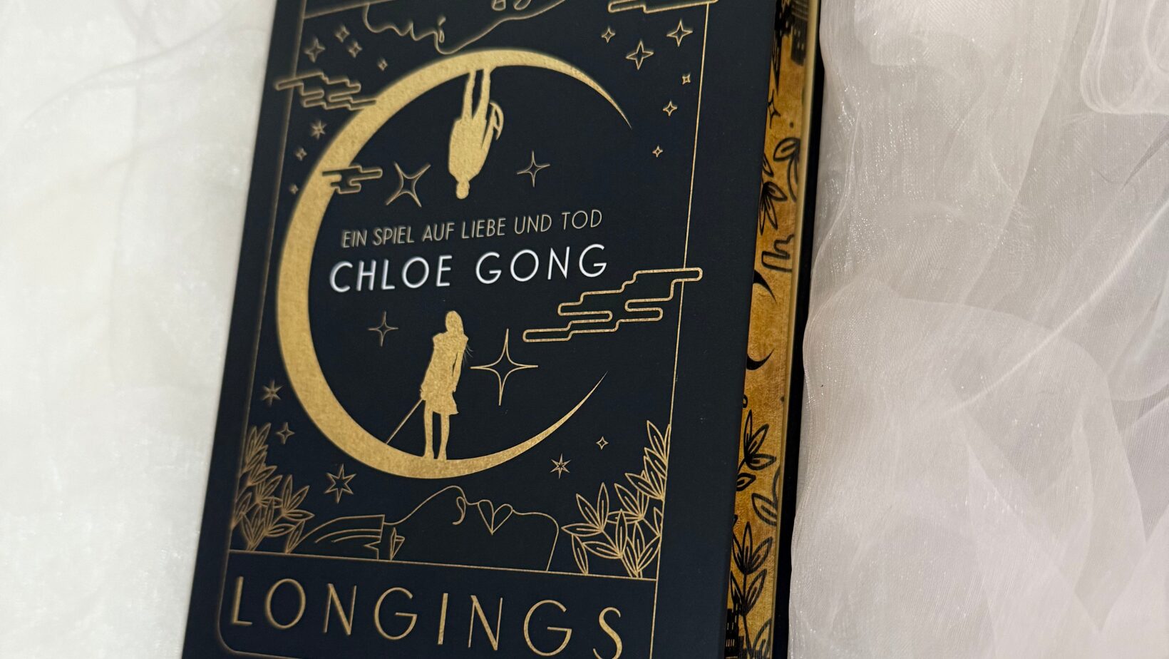 ein Buch mit einem schwarzen Einband und goldener Schrift. Der Titel des Buches lautet "IMMORTAL LONGINGS". Darunter steht "EIN SPIEL AUF LIEBE UND TOD" und der Name der Autorin "CHLOE GONG". Auf dem Cover ist ein großes, stilisiertes, goldenes Mondmotiv abgebildet, in dem zwei Figuren zu sehen sind – eine steht aufrecht und die andere hängt kopfüber. Der untere Teil des Covers zeigt eine Landschaft mit Pflanzen und Gebäuden. Unten rechts auf dem Cover ist das Logo von "Hobbit Presse Klett-Cotta" abgebildet. Das Buch liegt auf einem weißen, leicht transparenten Stoff.