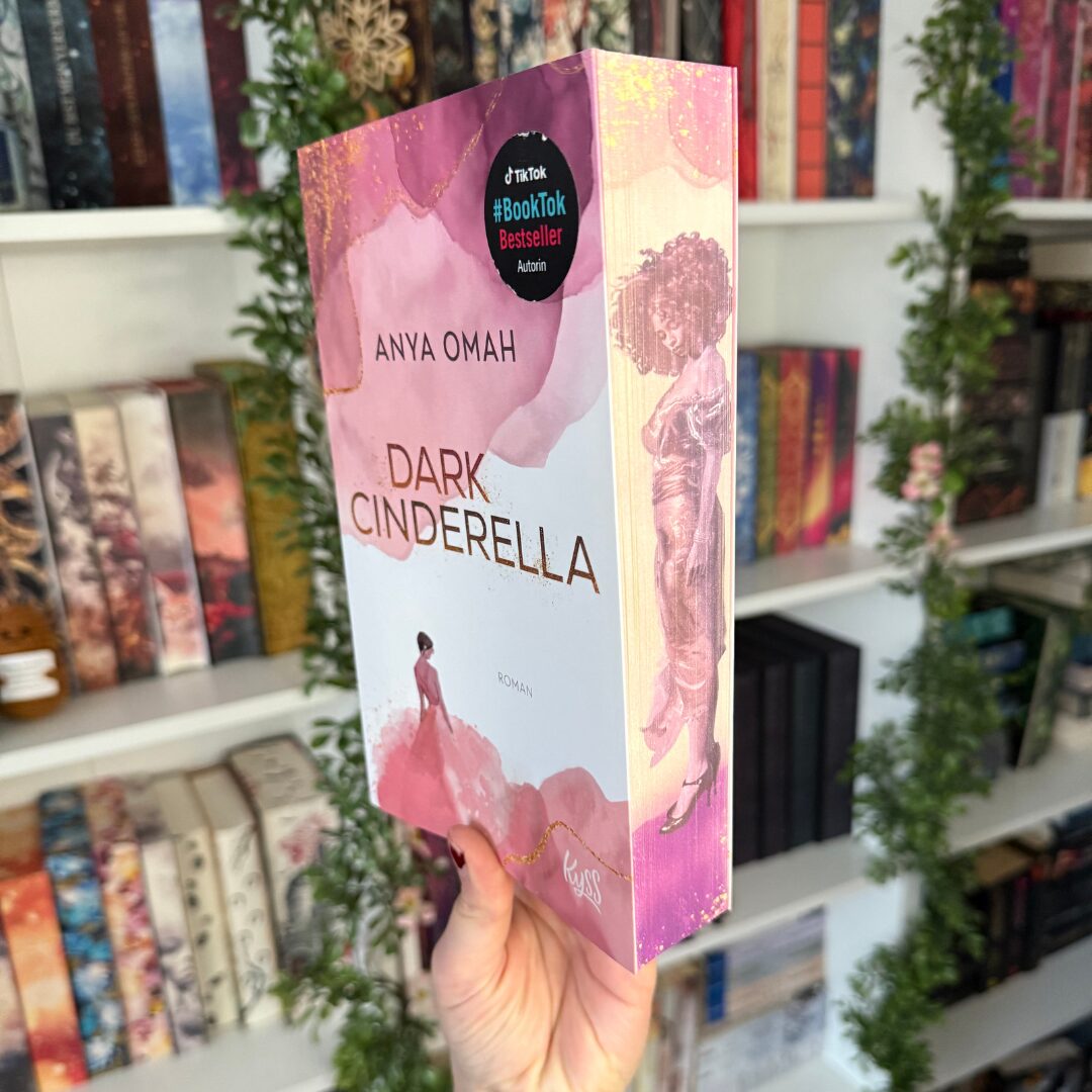 ein Buch, das in einer Hand gehalten wird. Der Titel des Buches ist "Dark Cinderella" und die Autorin ist Anya Omah. Auf dem Buchcover ist eine stilisierte Illustration einer Frau in einem rosa Kleid zu sehen. Oben auf dem Cover befindet sich ein runder Aufkleber mit der Aufschrift "#BookTok Bestseller Autorin". Im Hintergrund sind Bücherregale mit verschiedenen Büchern und grünen Pflanzen zu sehen. Das Buch hat einen rosa-weißen Farbverlauf und der Buchrücken zeigt eine weitere Illustration einer Frau in einem langen Kleid.