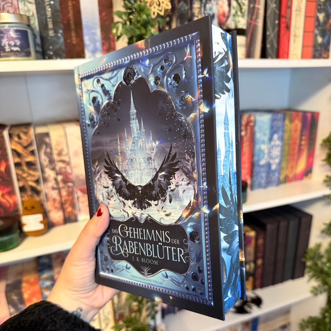 Eine Hand hält ein Buch mit dem Titel "Das Geheimnis der Rabenblüter" von J.K. Bloom. Das Cover zeigt ein düsteres, märchenhaftes Schloss im Hintergrund, umgeben von Raben und mystischen Elementen. Die Farben sind überwiegend in Blau- und Schwarztönen gehalten. Im Hintergrund sind Bücherregale mit verschiedenen Büchern und Dekorationen zu sehen, darunter Pflanzen und Kerzen. Die Atmosphäre wirkt magisch und geheimnisvoll.