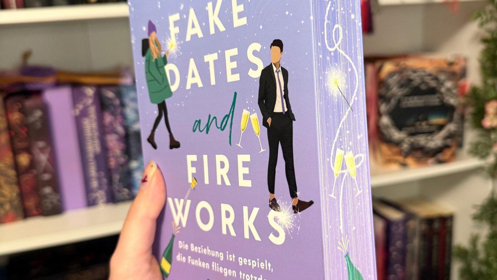 ein Buch, das in einer Hand gehalten wird. Der Titel des Buches lautet "Fake Dates and Fireworks" und die Autorin ist Kyra Groh. Das Cover ist in einem lila Farbton gestaltet und zeigt zwei stilisierte Figuren: eine Frau in einem grünen Mantel und einem lila Hut sowie einen Mann in einem Anzug. Zwischen ihnen sind zwei Sektgläser abgebildet. Auf dem Cover steht außerdem der Text: "Die Beziehung ist gespielt, die Funken fliegen trotzdem. Roman". Unten rechts ist das Logo des Verlags "Forever" zu sehen. Im Hintergrund sind Bücherregale mit verschiedenen Büchern und Dekorationen zu erkennen.