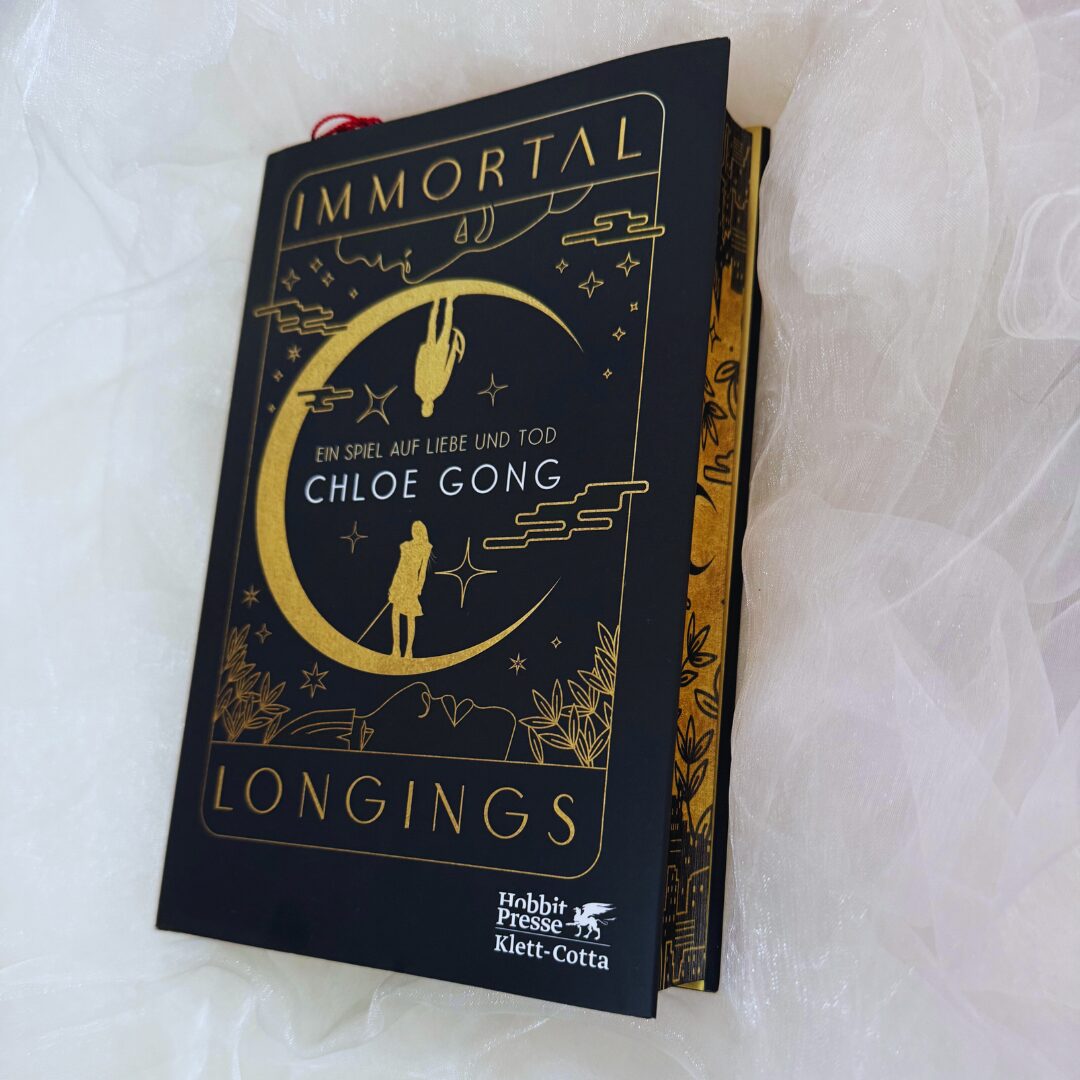 ein Buch mit einem schwarzen Einband und goldener Schrift. Der Titel des Buches lautet "IMMORTAL LONGINGS". Darunter steht "EIN SPIEL AUF LIEBE UND TOD" und der Name der Autorin "CHLOE GONG". Auf dem Cover ist ein großes, stilisiertes, goldenes Mondmotiv abgebildet, in dem zwei Figuren zu sehen sind – eine steht aufrecht und die andere hängt kopfüber. Der untere Teil des Covers zeigt eine Landschaft mit Pflanzen und Gebäuden. Unten rechts auf dem Cover ist das Logo von "Hobbit Presse Klett-Cotta" abgebildet. Das Buch liegt auf einem weißen, leicht transparenten Stoff.