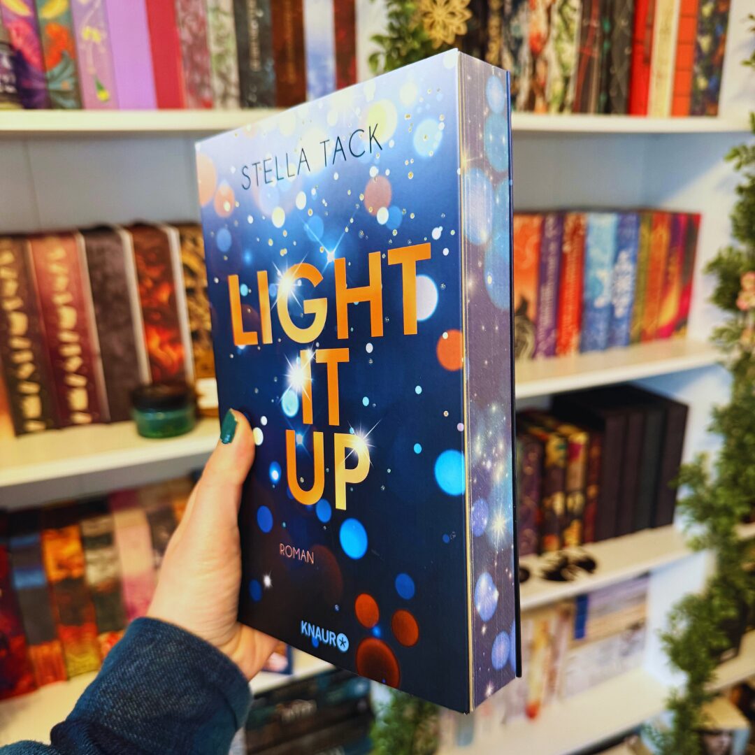 eine Hand hält ein Buch. Der Titel des Buches lautet "LIGHT IT UP" und der Autor ist Stella Tack. Auf dem Cover sind bunte Lichtpunkte in verschiedenen Farben wie Blau, Orange und Weiß zu sehen, die einen funkelnden Effekt erzeugen. Unten auf dem Cover steht "ROMAN" und das Logo des Verlags "KNAUR". Im Hintergrund sind Bücherregale mit verschiedenen Büchern zu erkennen, die in unterschiedlichen Farben und Größen angeordnet sind. Die Person hat grün lackierte Fingernägel und trägt einen blauen Pullover.