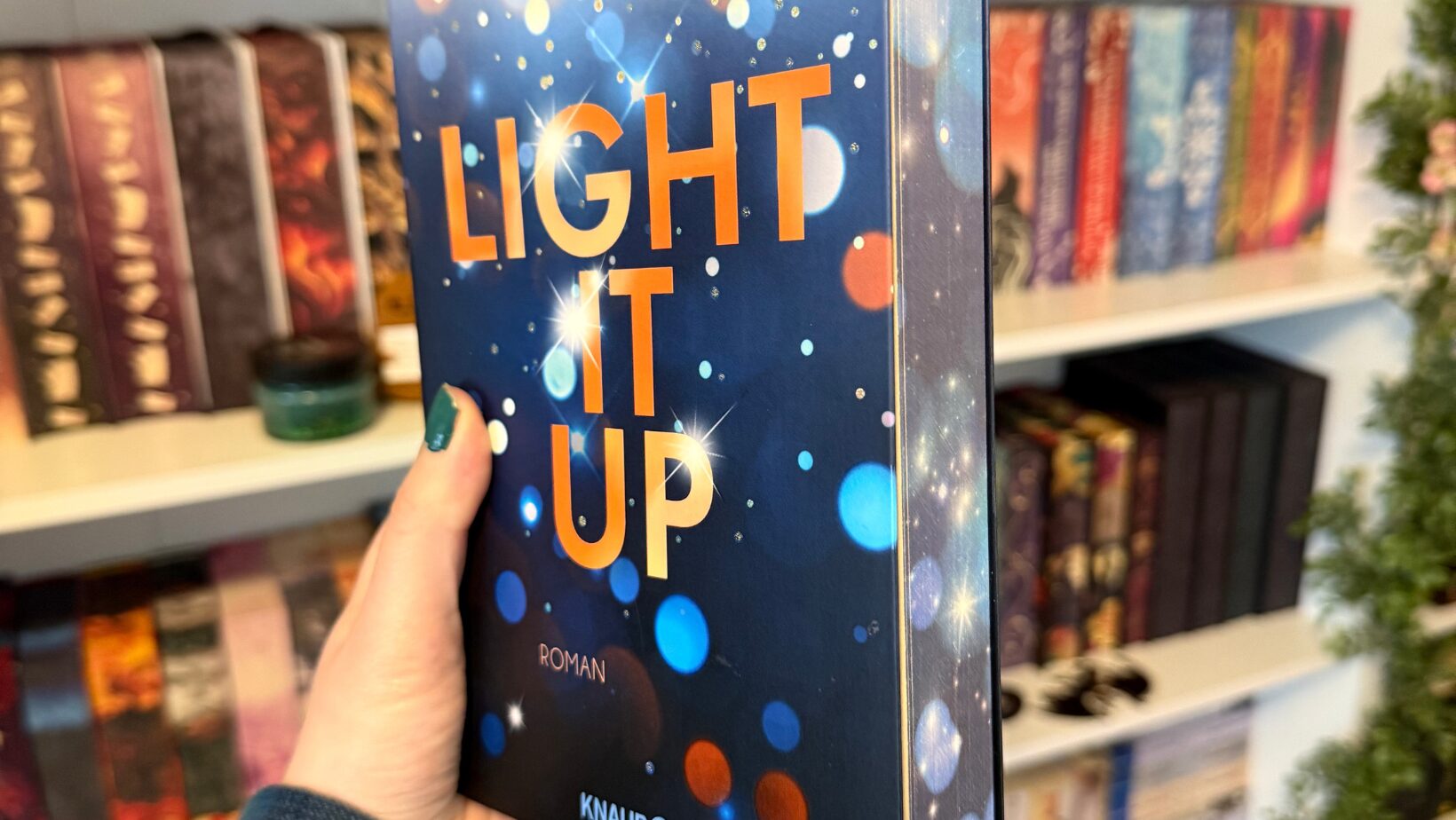 eine Hand hält ein Buch. Der Titel des Buches lautet "LIGHT IT UP" und der Autor ist Stella Tack. Auf dem Cover sind bunte Lichtpunkte in verschiedenen Farben wie Blau, Orange und Weiß zu sehen, die einen funkelnden Effekt erzeugen. Unten auf dem Cover steht "ROMAN" und das Logo des Verlags "KNAUR". Im Hintergrund sind Bücherregale mit verschiedenen Büchern zu erkennen, die in unterschiedlichen Farben und Größen angeordnet sind. Die Person hat grün lackierte Fingernägel und trägt einen blauen Pullover.