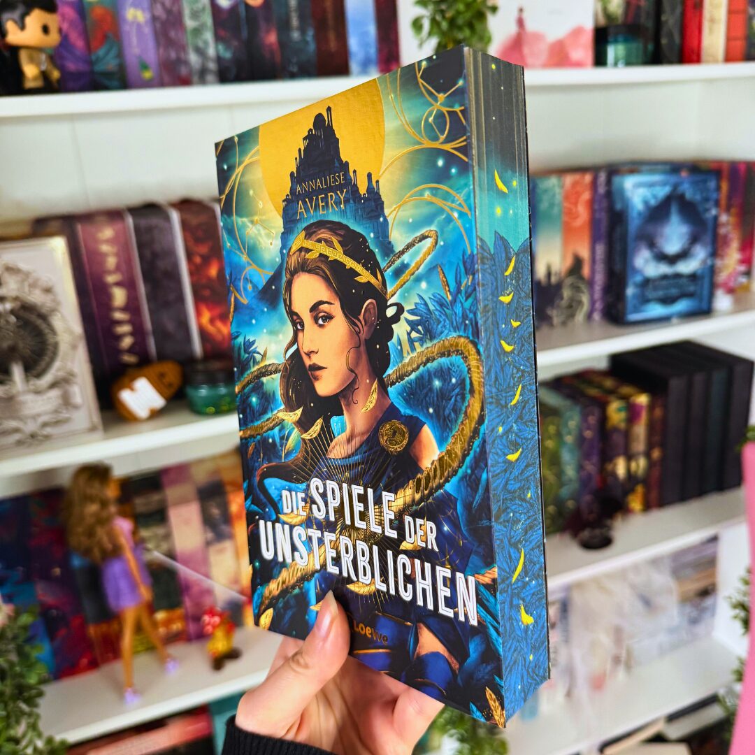 Ein Buch wird in der Hand gehalten. Der Titel des Buches lautet "Die Spiele der Unsterblichen" von Annaliese Avery. Das Cover zeigt eine Frau mit dunklem Haar, die von goldenen und blauen Elementen umgeben ist. Im Hintergrund ist eine mystische Landschaft mit einem großen Gebäude und einem leuchtenden Himmel zu sehen. Im Hintergrund des Bildes sind Bücherregale mit verschiedenen bunten Büchern und Figuren zu erkennen. Eine Barbie-Puppe in einem lila Kleid steht auf einem der Regale.