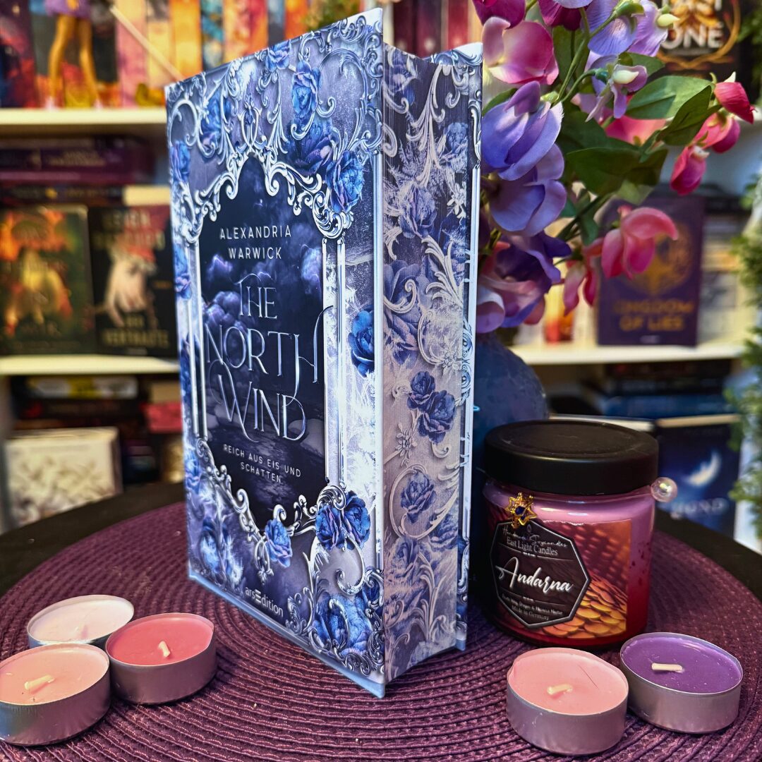 Buch mit verziertem Cover, mehrere Teelichter und eine Duftkerze. Im Mittelpunkt steht das Buch „The North Wind“ von Alexandria Warwick, das aufrecht auf einer lila Unterlage steht. Das Cover ist aufwendig gestaltet mit silbernen Ornamenten und blauen Rosen. Rechts daneben steht eine pink-lila Duftkerze mit schwarzem Deckel und dem Etikett „Andarna“. Um das Buch und die Kerze herum liegen fünf Teelichter in verschiedenen Farben: rosa, lila und weiß. Im Hintergrund sind weitere Bücherregale mit bunten Buchrücken und einige dekorative Blumen mit rosa und lila Blüten zu sehen. Die Szene wirkt gemütlich und farbenfroh, mit einem Fokus auf Buchliebhaber-Atmosphäre.