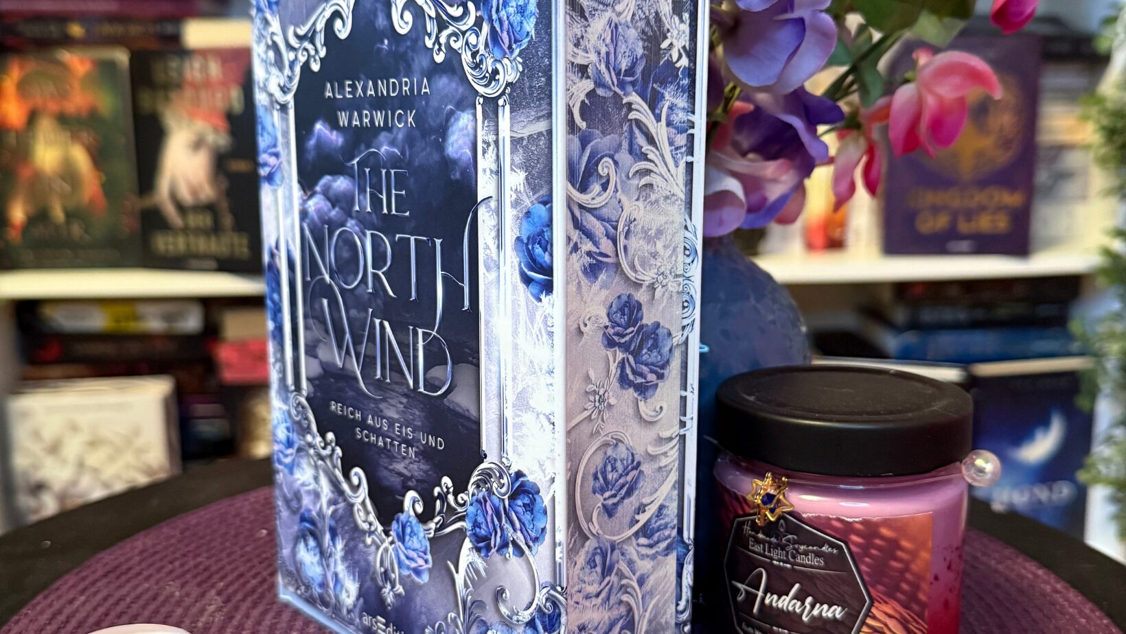 Buch mit verziertem Cover, mehrere Teelichter und eine Duftkerze. Im Mittelpunkt steht das Buch „The North Wind“ von Alexandria Warwick, das aufrecht auf einer lila Unterlage steht. Das Cover ist aufwendig gestaltet mit silbernen Ornamenten und blauen Rosen. Rechts daneben steht eine pink-lila Duftkerze mit schwarzem Deckel und dem Etikett „Andarna“. Um das Buch und die Kerze herum liegen fünf Teelichter in verschiedenen Farben: rosa, lila und weiß. Im Hintergrund sind weitere Bücherregale mit bunten Buchrücken und einige dekorative Blumen mit rosa und lila Blüten zu sehen. Die Szene wirkt gemütlich und farbenfroh, mit einem Fokus auf Buchliebhaber-Atmosphäre.
