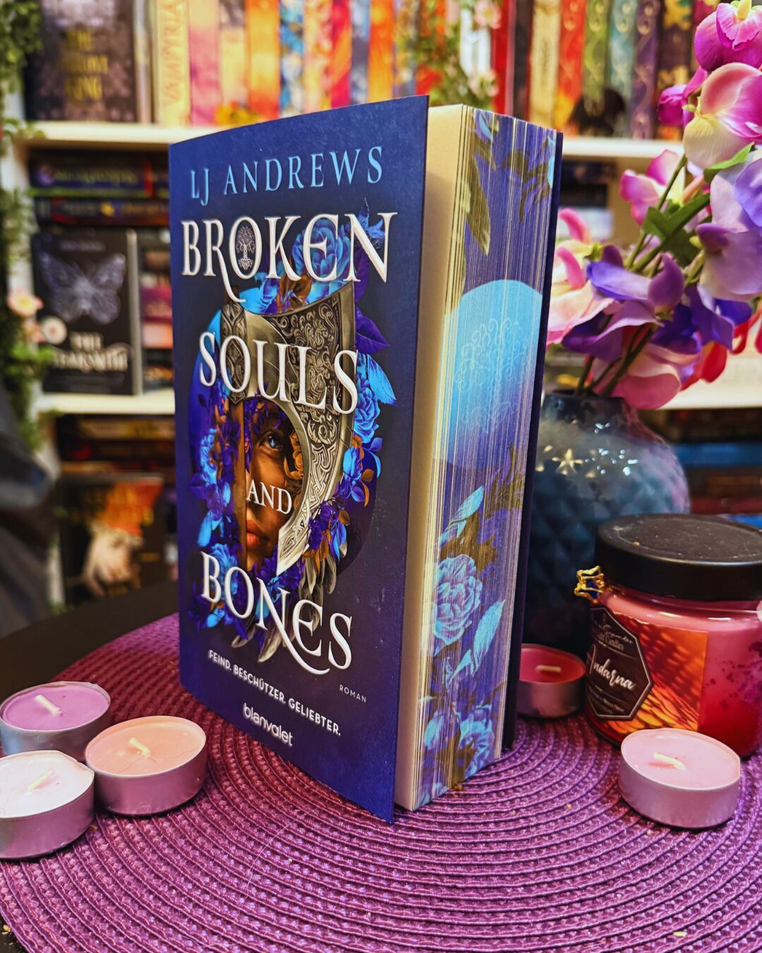 Ein aufrecht stehendes Buch mit dem Titel "Broken Souls and Bones" von LJ Andrews. Das Cover ist überwiegend blau und zeigt ein Gesicht, das zur Hälfte von einer verzierten silbernen Maske und blauen Blumen umrahmt ist. Unter dem Titel steht: "Feind. Beschützer. Geliebter. Roman" und das Verlagslogo "blanvalet". Das Buch steht auf einer lila, geflochtenen Unterlage. Rundherum liegen mehrere kleine Teelichter in Rosa- und Lilatönen. Rechts daneben steht eine rote Duftkerze mit schwarzem Deckel und goldfarbenem Etikett, auf dem "Habana" steht. Im Hintergrund ist eine blau-lila Vase mit künstlichen Blumen in Lila, Rosa und Weiß zu sehen. Dahinter befinden sich Bücherregale mit vielen bunten Buchrücken.