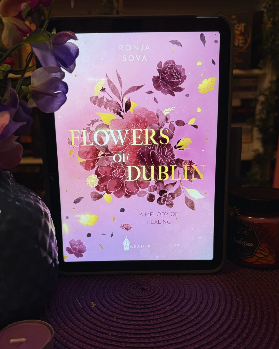 Ein Tablet mit einem Buchcover auf dem Bildschirm. Das Cover ist in Rosa- und Lilatönen gehalten und zeigt gezeichnete Blumen in verschiedenen Schattierungen von Lila, Rosa und Gelb. In der Mitte steht in großen, goldenen Buchstaben der Titel: "FLOWERS OF DUBLIN". Darüber steht der Name der Autorin: "RONJA SOVA". Unter dem Titel steht in kleinerer Schrift: "A MELODY OF HEALING". Unten ist das Logo des Verlags "WREADERS". Das Tablet steht auf einer lila, geflochtenen Unterlage. Links daneben ist eine lila Vase mit künstlichen Blumen und unten links ist eine lila Kerze zu sehen. Rechts steht ein Glas mit einem dunklen Deckel und einem Etikett. Die Atmosphäre wirkt gemütlich und einladend.