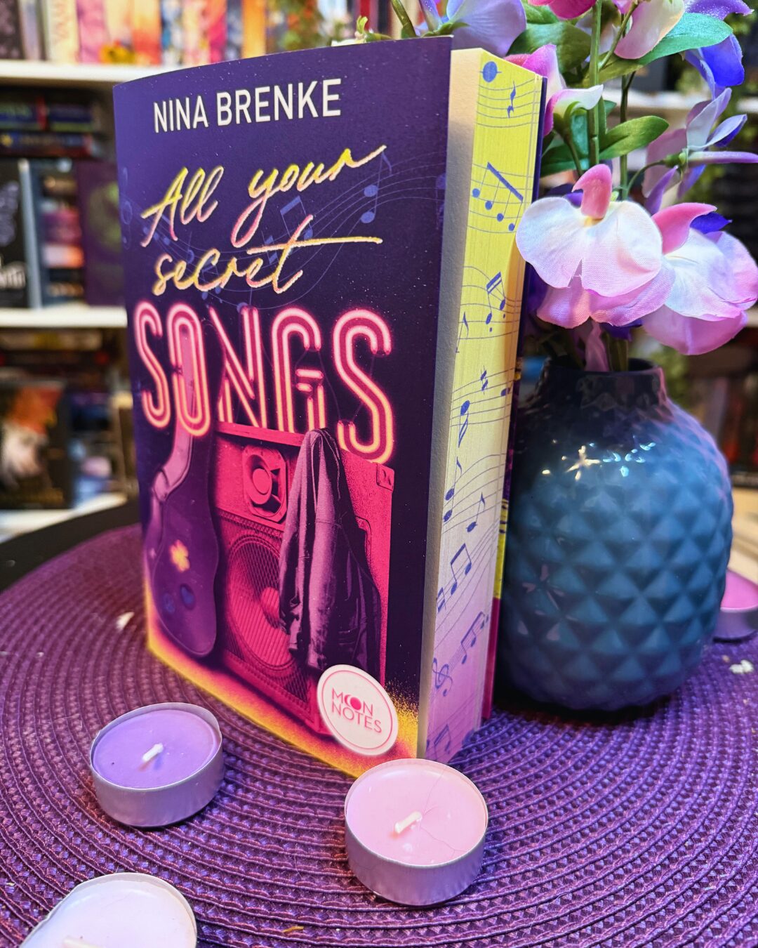 Buch mit dem Titel "All your secret SONGS" von Nina Brenke. Das Buch steht aufrecht auf einer lila geflochtenen Unterlage. Das Cover ist in Lila- und Pinktönen gehalten und zeigt eine Gitarre, einen Lautsprecher und eine Jacke. Im Hintergrund sind Notenlinien und Noten abgebildet. Neben dem Buch steht eine blaue Vase mit künstlichen rosa und lila Blüten. Vor dem Buch liegen drei kleine, lila Teelichter. Im Hintergrund sind unscharf weitere Bücherregale zu sehen.