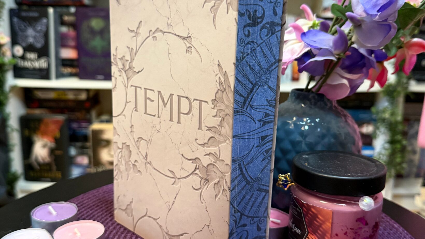 Ein Buch mit dem Titel "TEMPT" auf dem Einband. Das Buch steht aufrecht auf einer lila, geflochtenen Unterlage. Der Einband ist beige mit floralen Verzierungen, die wie in Stein gemeißelt aussehen, und der Buchrücken ist blau mit dekorativen Mustern. Um das Buch herum stehen mehrere kleine Teelichter in lila und rosa sowie ein Glas mit einer pinken Kerze und schwarzem Deckel. Im Hintergrund befindet sich eine dunkelblaue Vase mit künstlichen lila und rosa Blüten. Dahinter sieht man ein Regal voller bunter Bücher. Die Szene wirkt dekorativ und gemütlich, als wäre sie für ein Buchfoto oder eine Buchpräsentation arrangiert.