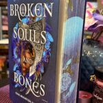 Ein aufrecht stehendes Buch mit dem Titel "Broken Souls and Bones" von LJ Andrews. Das Cover ist überwiegend blau und zeigt ein Gesicht, das zur Hälfte von einer verzierten silbernen Maske und blauen Blumen umrahmt ist. Unter dem Titel steht: "Feind. Beschützer. Geliebter. Roman" und das Verlagslogo "blanvalet". Das Buch steht auf einer lila, geflochtenen Unterlage. Rundherum liegen mehrere kleine Teelichter in Rosa- und Lilatönen. Rechts daneben steht eine rote Duftkerze mit schwarzem Deckel und goldfarbenem Etikett, auf dem "Habana" steht. Im Hintergrund ist eine blau-lila Vase mit künstlichen Blumen in Lila, Rosa und Weiß zu sehen. Dahinter befinden sich Bücherregale mit vielen bunten Buchrücken.