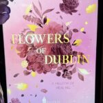 Ein Tablet mit einem Buchcover auf dem Bildschirm. Das Cover ist in Rosa- und Lilatönen gehalten und zeigt gezeichnete Blumen in verschiedenen Schattierungen von Lila, Rosa und Gelb. In der Mitte steht in großen, goldenen Buchstaben der Titel: "FLOWERS OF DUBLIN". Darüber steht der Name der Autorin: "RONJA SOVA". Unter dem Titel steht in kleinerer Schrift: "A MELODY OF HEALING". Unten ist das Logo des Verlags "WREADERS". Das Tablet steht auf einer lila, geflochtenen Unterlage. Links daneben ist eine lila Vase mit künstlichen Blumen und unten links ist eine lila Kerze zu sehen. Rechts steht ein Glas mit einem dunklen Deckel und einem Etikett. Die Atmosphäre wirkt gemütlich und einladend.