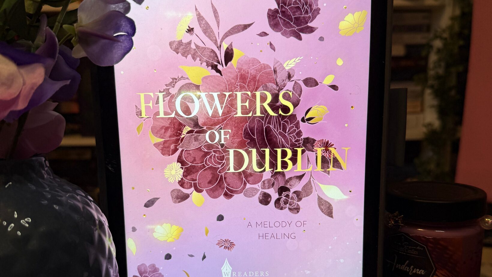Ein Tablet mit einem Buchcover auf dem Bildschirm. Das Cover ist in Rosa- und Lilatönen gehalten und zeigt gezeichnete Blumen in verschiedenen Schattierungen von Lila, Rosa und Gelb. In der Mitte steht in großen, goldenen Buchstaben der Titel: "FLOWERS OF DUBLIN". Darüber steht der Name der Autorin: "RONJA SOVA". Unter dem Titel steht in kleinerer Schrift: "A MELODY OF HEALING". Unten ist das Logo des Verlags "WREADERS". Das Tablet steht auf einer lila, geflochtenen Unterlage. Links daneben ist eine lila Vase mit künstlichen Blumen und unten links ist eine lila Kerze zu sehen. Rechts steht ein Glas mit einem dunklen Deckel und einem Etikett. Die Atmosphäre wirkt gemütlich und einladend.
