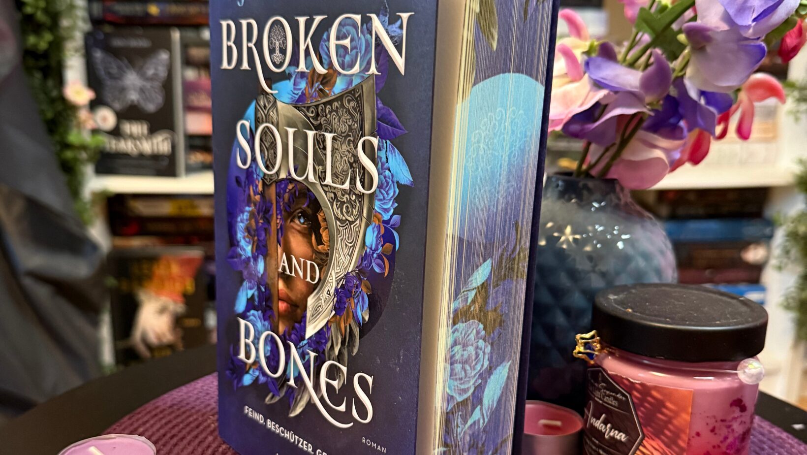 Ein aufrecht stehendes Buch mit dem Titel "Broken Souls and Bones" von LJ Andrews. Das Cover ist überwiegend blau und zeigt ein Gesicht, das zur Hälfte von einer verzierten silbernen Maske und blauen Blumen umrahmt ist. Unter dem Titel steht: "Feind. Beschützer. Geliebter. Roman" und das Verlagslogo "blanvalet". Das Buch steht auf einer lila, geflochtenen Unterlage. Rundherum liegen mehrere kleine Teelichter in Rosa- und Lilatönen. Rechts daneben steht eine rote Duftkerze mit schwarzem Deckel und goldfarbenem Etikett, auf dem "Habana" steht. Im Hintergrund ist eine blau-lila Vase mit künstlichen Blumen in Lila, Rosa und Weiß zu sehen. Dahinter befinden sich Bücherregale mit vielen bunten Buchrücken.