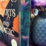 Buch mit farbig gestaltetem Buchschnitt, daneben eine blaue Vase mit künstlichen lila und rosa Blumen. Das Buch steht aufrecht auf einer lila, runden Platzmatte. Der Buchtitel lautet „Lessons of Hearts and Magic“ von Marion Meister. Im Hintergrund sind Bücherregale mit weiteren Büchern zu sehen. Die Buchseiten sind am Rand mit floralen Mustern und einem kleinen Vogel bedruckt. Die Szene wirkt farbenfroh und gemütlich, als wäre sie in einer Buchhandlung oder einem liebevoll dekorierten Lesezimmer aufgenommen worden.