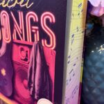 Buch mit dem Titel "All your secret SONGS" von Nina Brenke. Das Buch steht aufrecht auf einer lila geflochtenen Unterlage. Das Cover ist in Lila- und Pinktönen gehalten und zeigt eine Gitarre, einen Lautsprecher und eine Jacke. Im Hintergrund sind Notenlinien und Noten abgebildet. Neben dem Buch steht eine blaue Vase mit künstlichen rosa und lila Blüten. Vor dem Buch liegen drei kleine, lila Teelichter. Im Hintergrund sind unscharf weitere Bücherregale zu sehen.