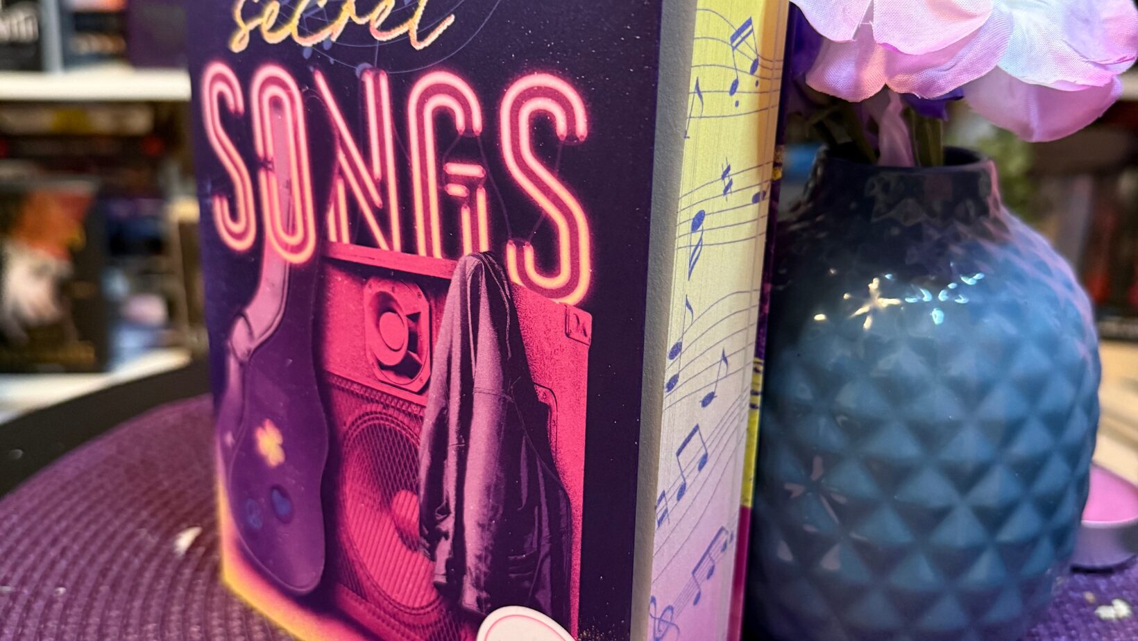 Buch mit dem Titel "All your secret SONGS" von Nina Brenke. Das Buch steht aufrecht auf einer lila geflochtenen Unterlage. Das Cover ist in Lila- und Pinktönen gehalten und zeigt eine Gitarre, einen Lautsprecher und eine Jacke. Im Hintergrund sind Notenlinien und Noten abgebildet. Neben dem Buch steht eine blaue Vase mit künstlichen rosa und lila Blüten. Vor dem Buch liegen drei kleine, lila Teelichter. Im Hintergrund sind unscharf weitere Bücherregale zu sehen.