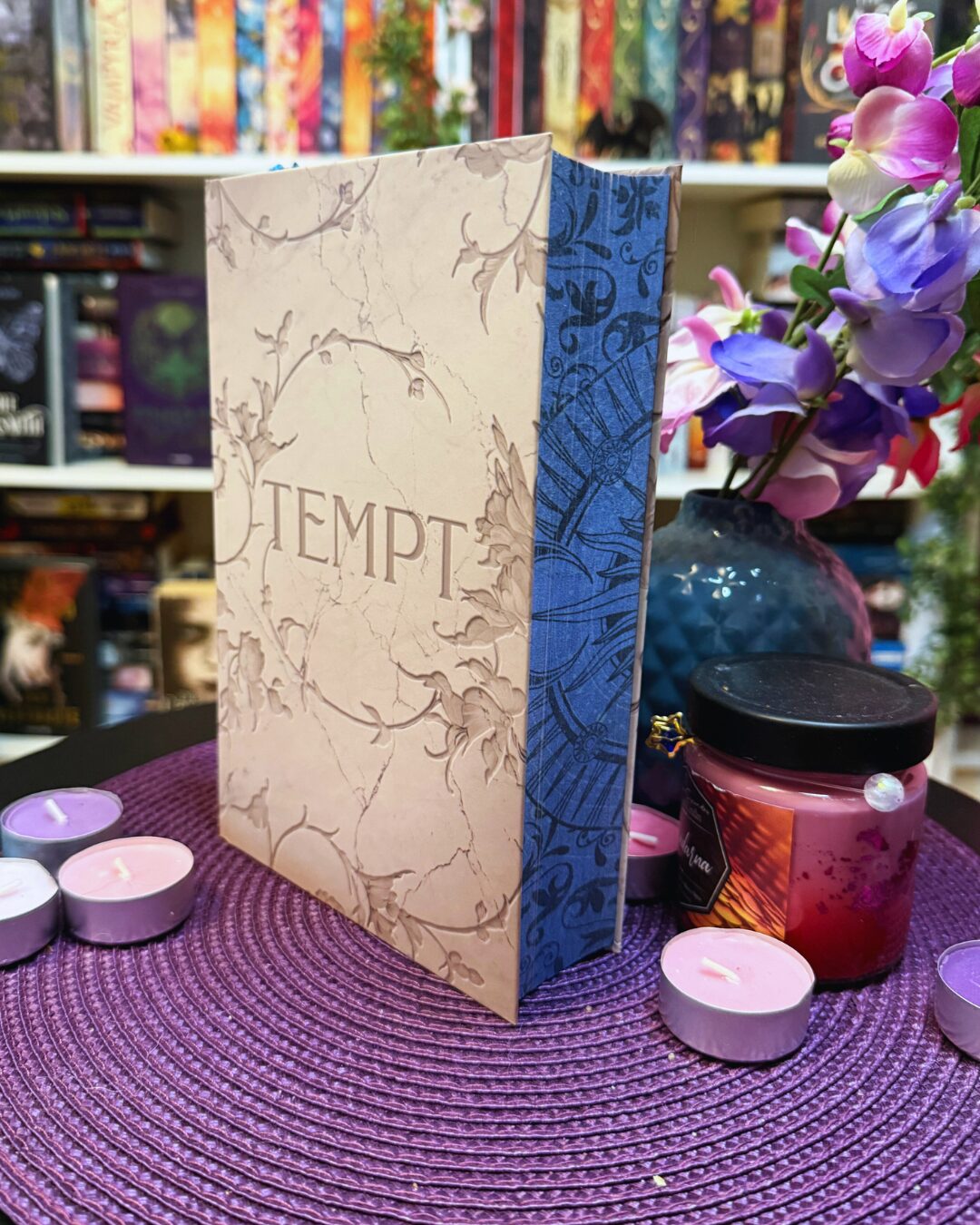 Ein Buch mit dem Titel "TEMPT" auf dem Einband. Das Buch steht aufrecht auf einer lila, geflochtenen Unterlage. Der Einband ist beige mit floralen Verzierungen, die wie in Stein gemeißelt aussehen, und der Buchrücken ist blau mit dekorativen Mustern. Um das Buch herum stehen mehrere kleine Teelichter in lila und rosa sowie ein Glas mit einer pinken Kerze und schwarzem Deckel. Im Hintergrund befindet sich eine dunkelblaue Vase mit künstlichen lila und rosa Blüten. Dahinter sieht man ein Regal voller bunter Bücher. Die Szene wirkt dekorativ und gemütlich, als wäre sie für ein Buchfoto oder eine Buchpräsentation arrangiert.