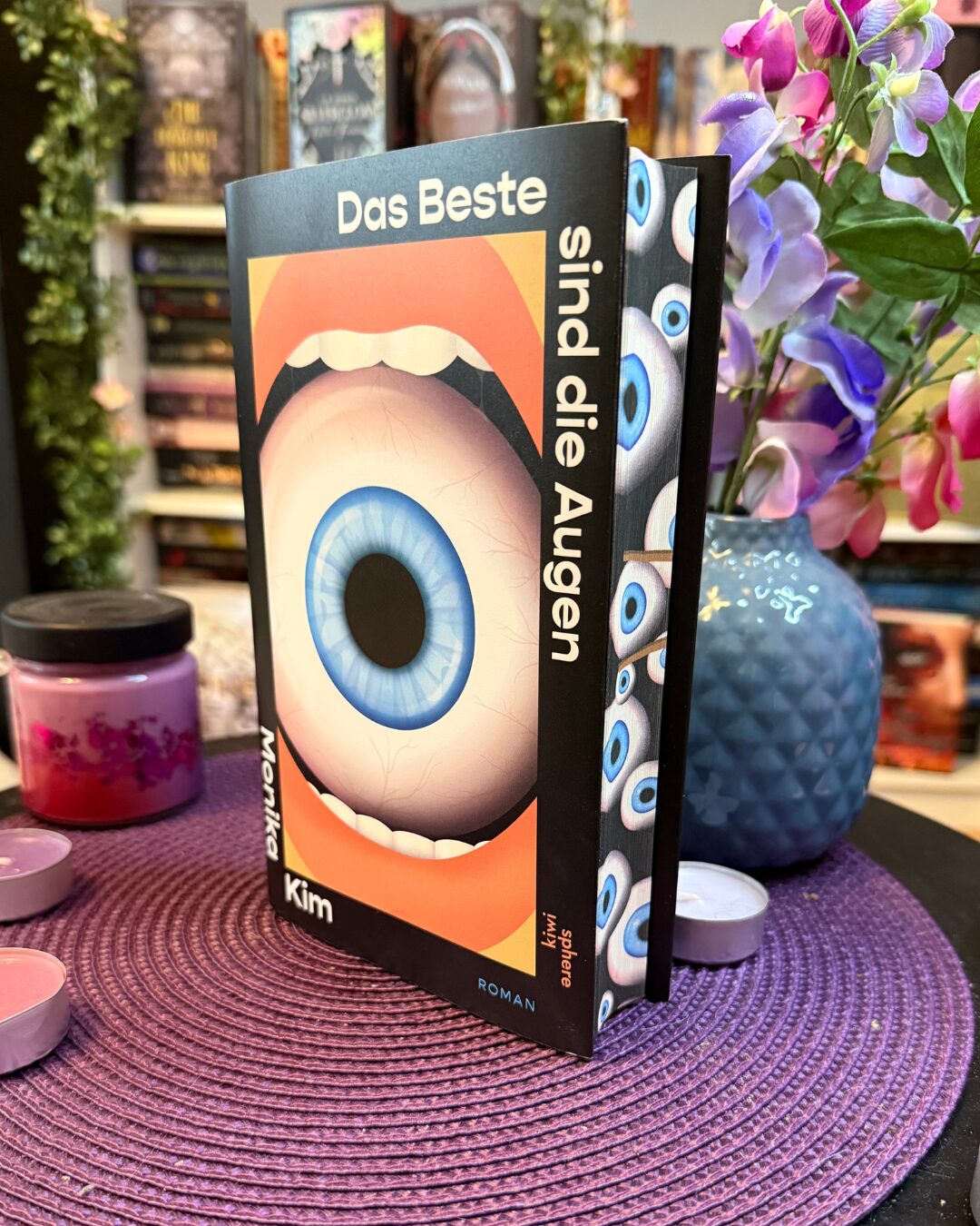 Buch mit auffälligem Cover: Das Buch steht aufrecht auf einer lila, geflochtenen Unterlage. Das Cover zeigt einen großen, blauen Augapfel, der in einem offenen Mund mit weißen Zähnen platziert ist. Der Titel lautet „Das Beste sind die Augen“ und der Autorenname ist Monika Kim. Am Buchschnitt sind viele kleine, blaue Augen abgebildet. Im Hintergrund stehen weitere Bücher in einem Regal, daneben sieht man eine blaue Vase mit lila und pinken Blumen sowie einige rosafarbene Teelichter und eine Kerze. Die Szene wirkt farbenfroh und kreativ.