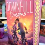 Buch mit dem Titel "Downhill Dreams" von Nadine Schjoer, aufrecht stehend auf einem fliederfarbenen, geflochtenen Platzset. Das Buch zeigt auf dem Cover zwei Personen mit Fahrrädern, die sich nahe stehen, vor einer Sonnenuntergangskulisse und Bergen im Hintergrund. Das Buch hat pinke Seitenkanten mit einem gelben Muster. Vor dem Buch stehen mehrere kleine, rosa Teelichter und eine pinke Duftkerze in einem Glas. Im Hintergrund sieht man ein Regal mit bunten Buchrücken und einige grüne künstliche Ranken, die von den Regalen hängen. Die Atmosphäre wirkt gemütlich und liebevoll dekoriert.