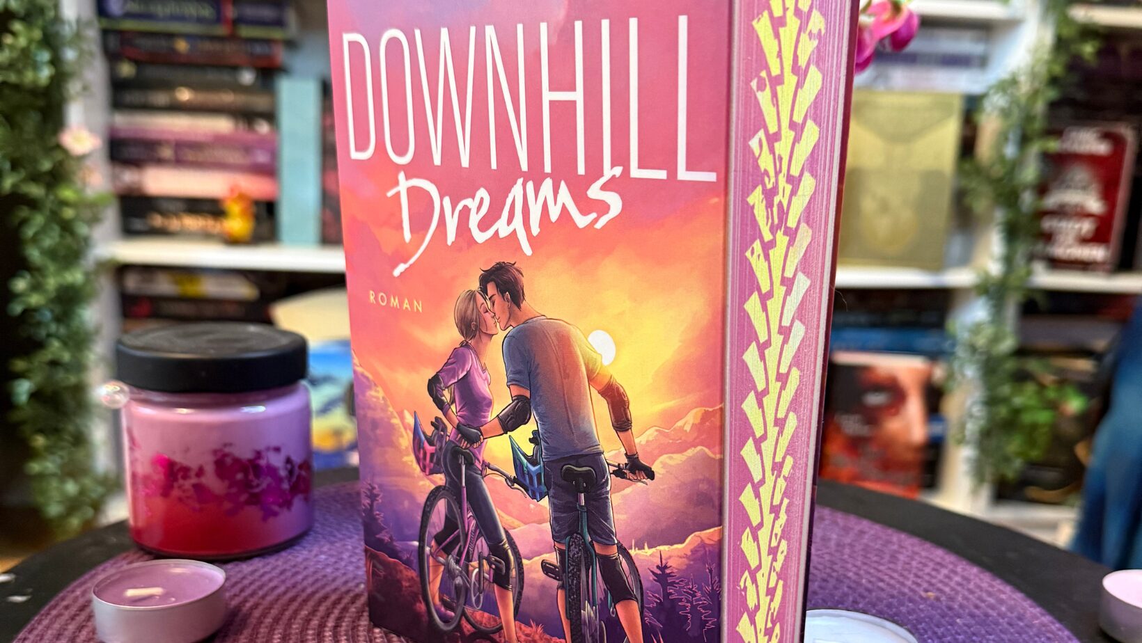 Buch mit dem Titel "Downhill Dreams" von Nadine Schjoer, aufrecht stehend auf einem fliederfarbenen, geflochtenen Platzset. Das Buch zeigt auf dem Cover zwei Personen mit Fahrrädern, die sich nahe stehen, vor einer Sonnenuntergangskulisse und Bergen im Hintergrund. Das Buch hat pinke Seitenkanten mit einem gelben Muster. Vor dem Buch stehen mehrere kleine, rosa Teelichter und eine pinke Duftkerze in einem Glas. Im Hintergrund sieht man ein Regal mit bunten Buchrücken und einige grüne künstliche Ranken, die von den Regalen hängen. Die Atmosphäre wirkt gemütlich und liebevoll dekoriert.