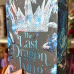 Nahaufnahme eines Buches mit dem Titel „The Last Dragon King“ von Leia Stone. Das Buchcover zeigt eine große, kristallene Krone sowie leuchtende, blaue und feurige Elemente. Das Buch wird von einer Hand mit schwarzem Ärmel gehalten. Im Hintergrund sind mehrere bunt gestaltete Bücherregale mit weiteren Fantasy-Büchern, Dekorationen wie Pflanzenranken und einem kleinen Funko-Pop zu sehen. Die Szene wirkt wie ein gemütlicher Buchliebhaber-Bereich.