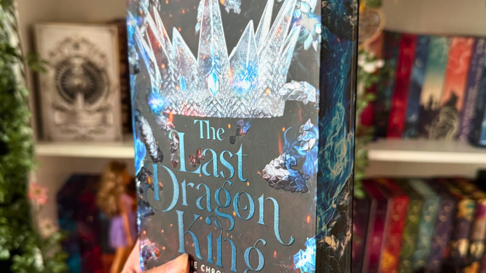 Nahaufnahme eines Buches mit dem Titel „The Last Dragon King“ von Leia Stone. Das Buchcover zeigt eine große, kristallene Krone sowie leuchtende, blaue und feurige Elemente. Das Buch wird von einer Hand mit schwarzem Ärmel gehalten. Im Hintergrund sind mehrere bunt gestaltete Bücherregale mit weiteren Fantasy-Büchern, Dekorationen wie Pflanzenranken und einem kleinen Funko-Pop zu sehen. Die Szene wirkt wie ein gemütlicher Buchliebhaber-Bereich.